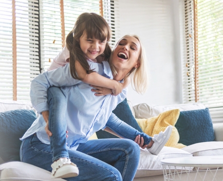 Happy Beautiful Mother carrying or piggyback her little daughter laughing playing and having fun together on sofa at home.